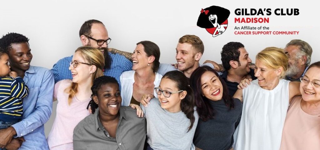 The image shows a group of smiling people posing for a photo along with a logo for Gilda's Club Madison (an affiliate of the Cancer Support Community). The group consists of individuals of various ages, races, and genders. The setting is indoors against a white wall.