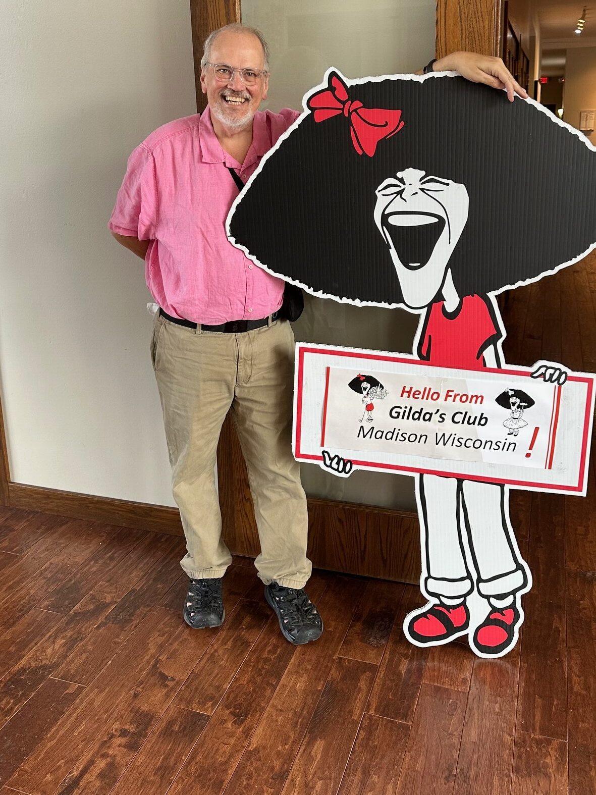 Ben standing next to a life size cutout cartoon of Gilda who is holding a sign that reads "Hello from Gilda's Club Madison Wisconsin!"